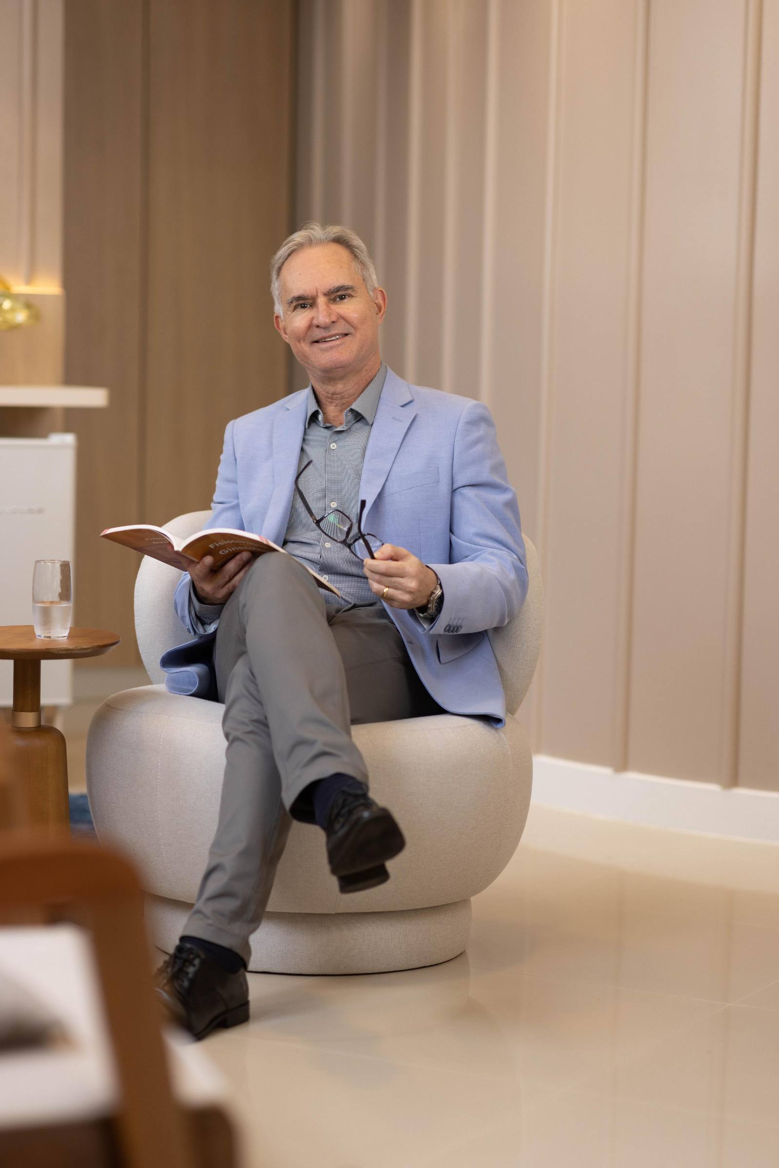Dr. Evandro Vanti sentado em uma poltrona clara, de blazer azul, com um livro aberto e os óculos na mão
